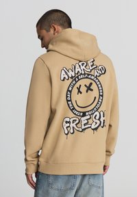 Felpa color beige con una grande grafica di un sorriso nero sul retro, che presenta il testo "AWARE AND FRESH" in un font giocoso. Tessuto dalla consistenza liscia.