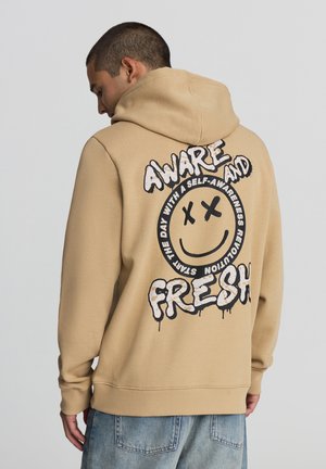 Felpa color beige con una grande grafica di un sorriso nero sul retro, che presenta il testo "AWARE AND FRESH" in un font giocoso. Tessuto dalla consistenza liscia.