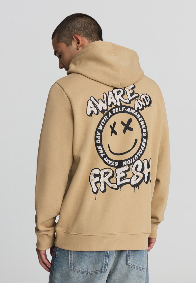 Felpa color beige con una grande grafica di un sorriso nero sul retro, che presenta il testo "AWARE AND FRESH" in un font giocoso. Tessuto dalla consistenza liscia.
