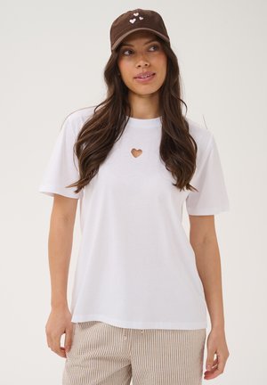 Femme aux longs cheveux bruns portant une casquette marron avec des cœurs, un t-shirt blanc avec une petite ouverture en forme de cœur, et un pantalon beige à rayures.