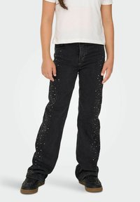 Jeans de mezclilla negros con un corte recto, decorados con tachuelas plateadas a lo largo de las costuras laterales. Se combinan con una camisa blanca de manga corta y zapatillas negras.