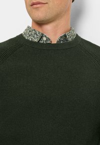 Dunkelgrüner gerippter Pullover mit Rundhalsausschnitt, darüber ein leuchtendes, floral gemustertes Hemd mit Knopfleiste und Kragen.