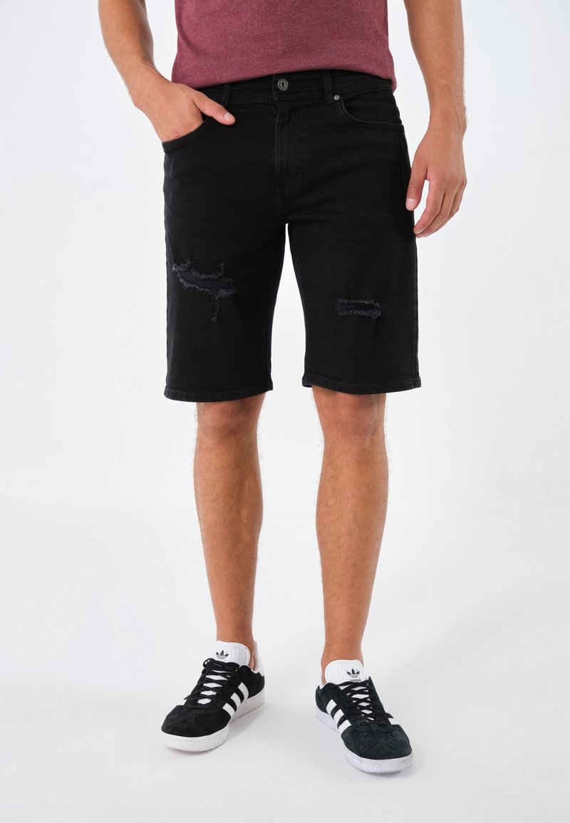Pantalones cortos de mezclilla negra, hasta la rodilla, con desgastes y rotos. Se llevan con zapatillas negras y blancas. El modelo está de pie de manera casual con una mano en el bolsillo.