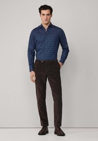 Camisa a cuadros azul marino con cuello abotonado, combinada con pantalones de pana marrón oscuro y botas negras con cordones. Fondo minimalista.