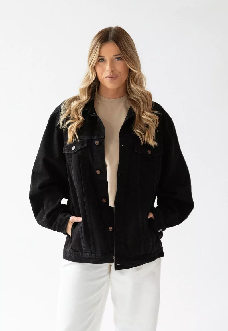 Czarna kurtka dżinsowa o luźnym kroju, z zapięciem na guziki, kieszeniami na piersi oraz kieszeniami bocznymi; zestawiona z beżową bluzką i białymi spodniami.