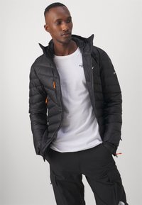 Giacca a vento nera con design trapuntato, dotata di zip arancioni e cappuccio, indossata sopra una t-shirt bianca e pantaloni cargo neri.