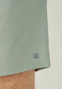 Jasnozielone spodenki sportowe wykonane z teksturowanej tkaniny z małą szaro-białą naszywką przy dolnym szwie, prezentujące prosty i funkcjonalny design.