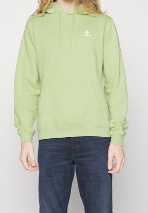 Osoba ubrana w jasnobrzozowy zielony kaptur Converse z przednią kieszenią i ciemnoniebieskie dżinsy, stojąca na jednolitym białym tle.