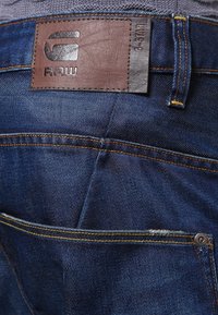 Jeans de denim en azul oscuro con parche de cuero marrón. Presentan costuras visibles, pasadores de cinturón y un pequeño botón de metal en el bolsillo.