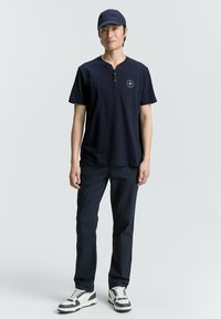 Jūrinės mėlynos spalvos polo marškiniai su trumpomis rankovėmis ir henley tipo apykakle, papuošti siuvinėtu logotipu, derinami su jūrinės mėlynos spalvos kelnėmis ir juodai baltais sportbačiais. Kasdienis aprangos stilius.