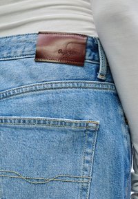 Pantalones vaqueros de mezclilla azul claro con una etiqueta de cuero marrón en la cinturilla. Presentan dos bolsillos traseros y detalles de costuras visibles.