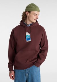 Vinchansad mörkröd hoodie med framlapp och en vit och blå grafisk etikett, kombinerad med ljusblå denimbyxor och en grön stickad mössa.