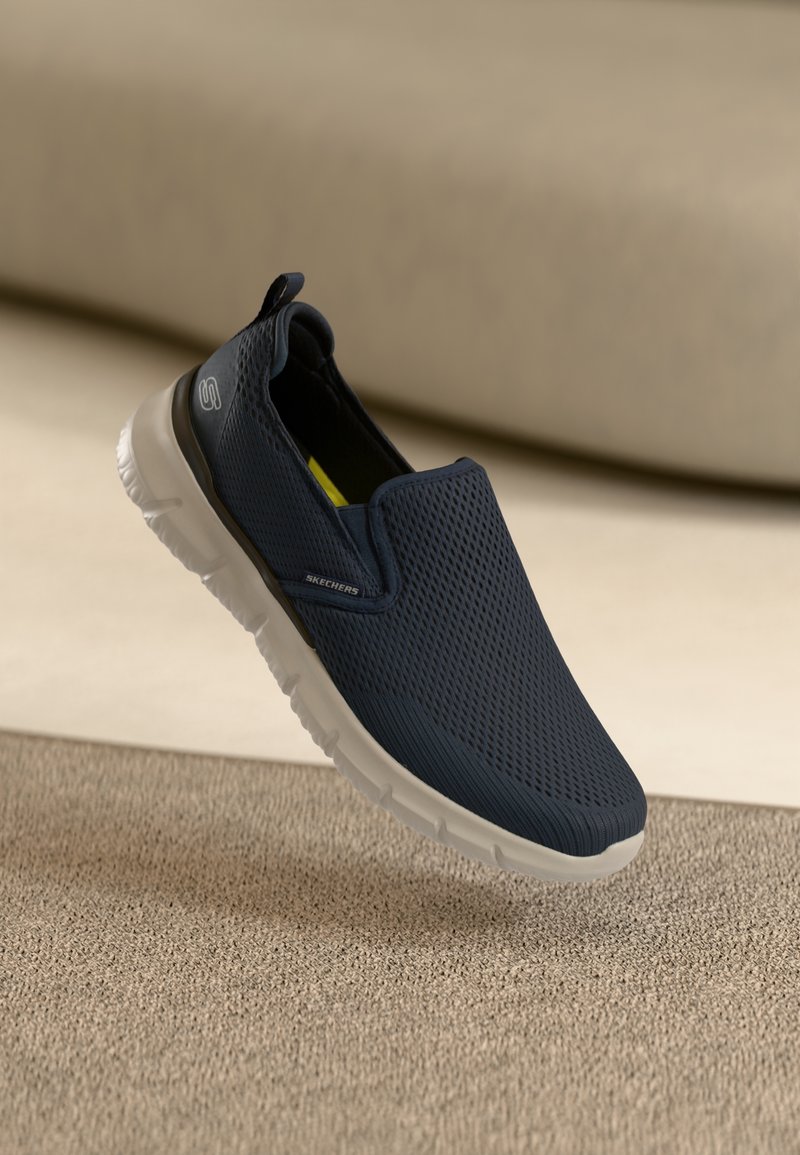 Baskets Skechers bleu marine à enfiler avec semelle blanche, dessus en mesh respirant et languette sur une surface texturée neutre.