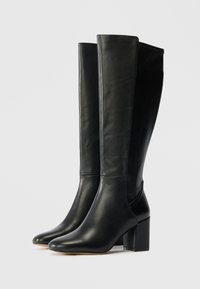 Juodos odinės kelio ilgio botinės su lygiu paviršiumi, smailu priekiu ir blokuotu kulnu. Išsiskiria minimalia siūlių danga ir elegantiška siluetu.