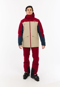 Skijacke in Beige,Weinrot und Marine, mit hohem Kragen, Reißverschluss vorne und mehreren Taschen. Kombiniert mit weinroten Skihosen und schwarzen Stiefeln.