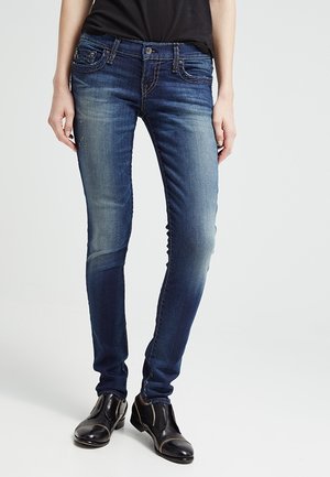 Person iført falmede blå slim fit jeans, sorte ankelstøvler med lynlåse og en sort top, stående mod en ensfarvet lys baggrund.