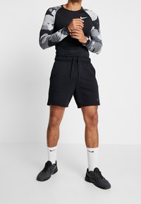 Czarne szorty sportowe z elastycznym pasem, połączone z czarną bluzą z długim rękawem z szarymi abstrakcyjnymi wzorami. Czarne sneakersy i białe skarpetki.