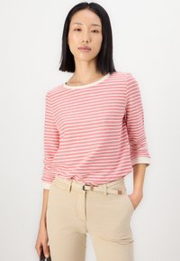Top a maniche lunghe a righe corallo e bianche con scollo rotondo e polsini color crema, abbinato a pantaloni beige attillati e una cintura intrecciata.