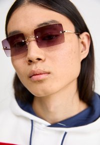 Rectangular purple sunglasses with wire frames and decorative hardware. Worn with a white hoodie featuring red and navy design accents.