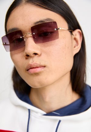 Téglalap alakú, lila napszemüveg fémből készült kerettel és díszes elemekkel. Fehér kapucnis pulóvert visel, melynek piros és tengerészkék dizájn díszítése van.