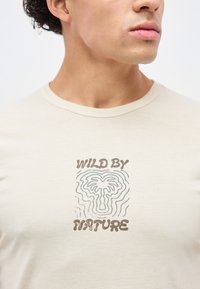 Άνδρας φοράει μπεζ t-shirt με το κείμενο "WILD BY NATURE" και αφηρημένο φύλλο σχεδιασμένο στο κέντρο, στραμμένος ελαφρώς προς τα δεξιά.