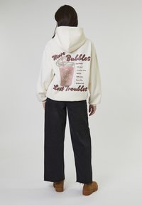 Persoon in een crèmekleurige hoodie met een roze bubble tea-illustratie en de tekst "Meer Bubbels Minder Problemen", zwarte wijde broek en bruine laarzen, staand met de rug naar ons toe.