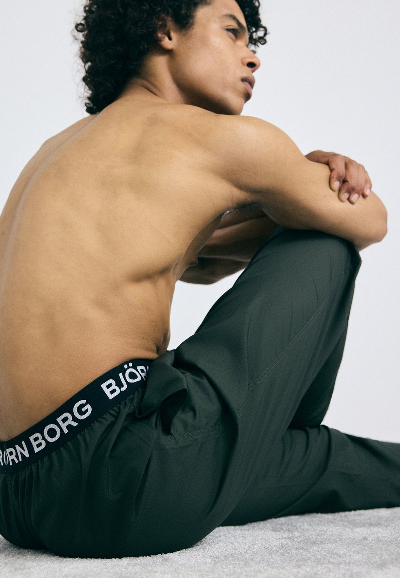 Calças leves de cor verde-escuro com uma cintura elástica preta que apresenta texto branco. Modelo sentado de lado sobre uma superfície texturizada, costas nuas visíveis.