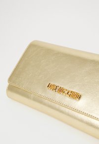 Portefeuille en cuir métallique doré avec plaque logo "LOVE MOSCHINO" dorée sur le rabat avant, sur fond blanc.