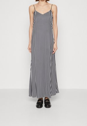 Woman wearing a black and white vertical striped sleeveless maxi dress with thin straps and black loafers, standing against a plain background.