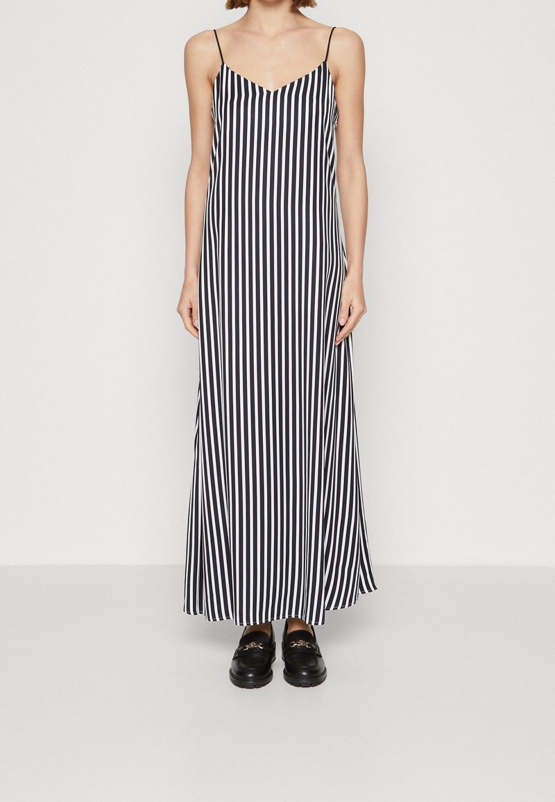 Femme portant une robe maxi sans manches à rayures verticales noires et blanches avec des bretelles fines et des mocassins noirs, debout devant un fond uni.