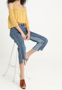 Yellow off-the-shoulder top with lace trim, paired with blue split-hem jeans. Model wears brown open-toe heeled mules. White background.