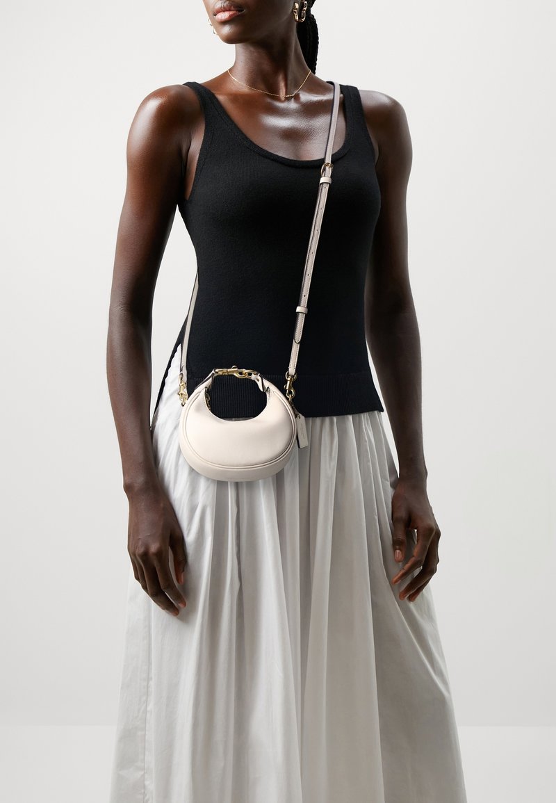 Femme portant un haut noir sans manches et une jupe blanche porte un petit sac bandoulière blanc avec des ferrures dorées.
