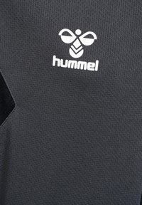 Hvid Hummel-logo med stiliseret bi og tekst på mørkegråt åndbart sportsstof.