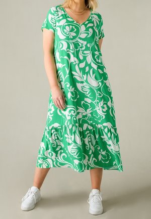 Vrouw draagt een groen-wit bloemenmidi-jurk met korte mouwen en een gelaagde zoom, gecombineerd met witte sneakers, staand tegen een effen achtergrond.