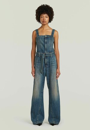 Femme aux cheveux bouclés portant une combinaison en denim bleu sans manches avec une ceinture, se tenant devant un fond clair uni.