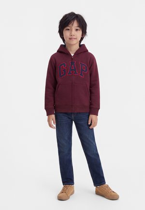 Enfant aux cheveux foncés portant un sweat à capuche bordeaux GAP, un jean bleu et des baskets beige, debout devant un fond blanc uni.