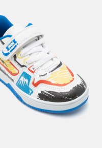 Sneaker blanc avec des motifs abstraits multicolores en rouge, jaune, bleu et noir ; présente une accentuation bleue, une sangle Velcro et une semelle texturée.