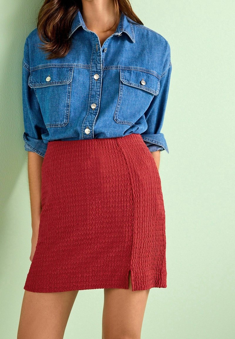 La camicia in denim presenta tasche sul petto e chiusure a bottone, abbinata a una gonna rossa strutturata con spacco laterale e design aderente.