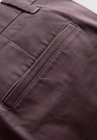 Dunkelbrauner Stoff mit einer glatten Textur, der eine diagonale Naht und eine einfache, schmale Tasche auf der rechten Seite aufweist.