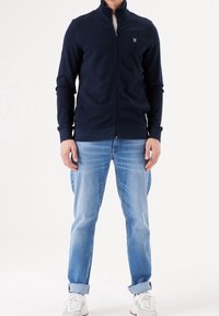 Giacca zip-up blu navy in un materiale morbido, con colletto alto e polsini a coste, abbinata a jeans azzurri e sneakers bianche.