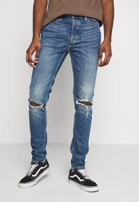 Hombre vestido con jeans azules rasgados, zapatillas negras y blancas, y una camisa de rayas marrón, de pie contra un fondo blanco liso.