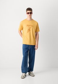 Gul cotton t-skjorte med trykket fugledesign og "Makia"-tekst, sammen med løse blå denimjeans og grå sneakers med brune detaljer.