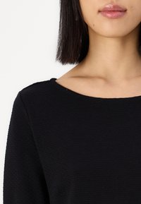 Svart teksturert topp med rund halslinning, som har en jevn finish og korte ermer, med et subtilt ribbet mønster.