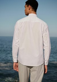 White striped long-sleeve shirt, buttoned cuffs, slight sheen, paired with light gray striped trousers, outdoors near water.