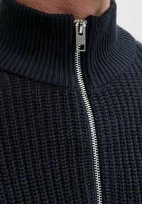 Ciemnoniebieski sweter wykonany na drutach z wysokim kołnierzem, z ozdobnym srebrnym zamkiem i teksturowanym wzorem. Materiał wydaje się miękki i ciepły.