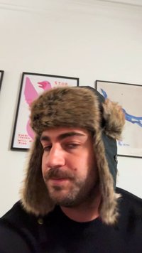 Černá letecká čepice s umělým kožíškem na uších. Kožíšek je béžový a hnědý, což kontrastuje s tmavým materiálem. Obsahuje knoflík po straně.