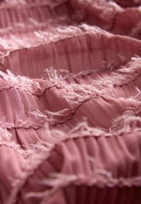 Tekstureret pink stof med frynsede kanter, der har et plisseret design. Blødt materiale med en let skinnende overflade og synlige mønstervariationer.