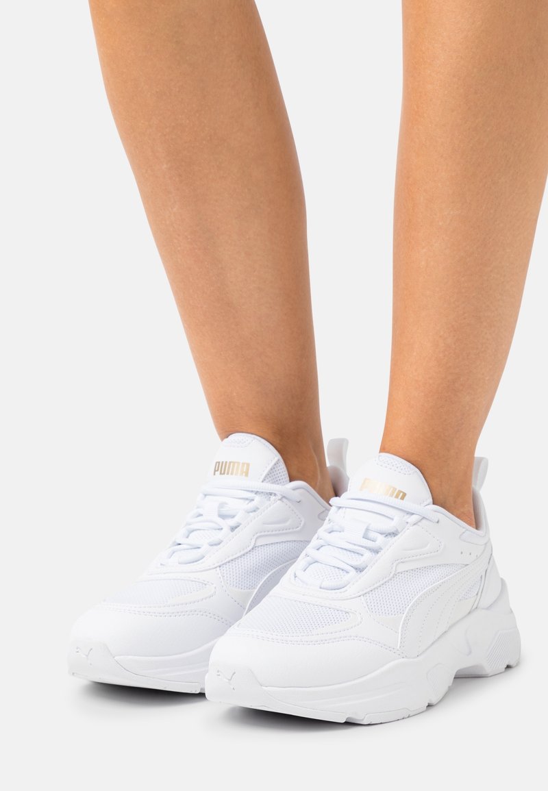Jambes portant des baskets blanches Puma avec des détails en mesh et cuir, lacées, debout côte à côte sur un fond clair.