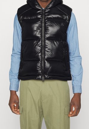 Person trägt glänzende schwarze Steppweste mit Kapuze über hellblauem Langarmshirt und olivgrüner Cargohose, Vorderansicht.