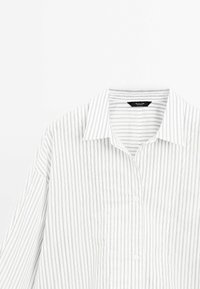 Camicia bianca a maniche lunghe con bottoni, a righe verticali nere sottili e colletto classico, poggiata su uno sfondo bianco.